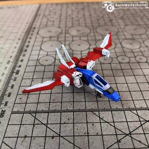 May include: AeroModelBuild model airplane. The model is a small, detailed aircraft with a red, white, and blue color scheme. The wings and tail are red and white, while the fuselage is blue. The model is on a grid surface.