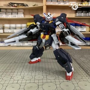 Puede incluir: Modelo detallado de un robot Gundam, con una combinaci&oacute;n de colores azul marino, blanco, rojo y amarillo. El robot tiene grandes alas y est&aacute; posado sobre una superficie cuadriculada. El fondo incluye estantes con botes de pintura y suministros de modelismo.