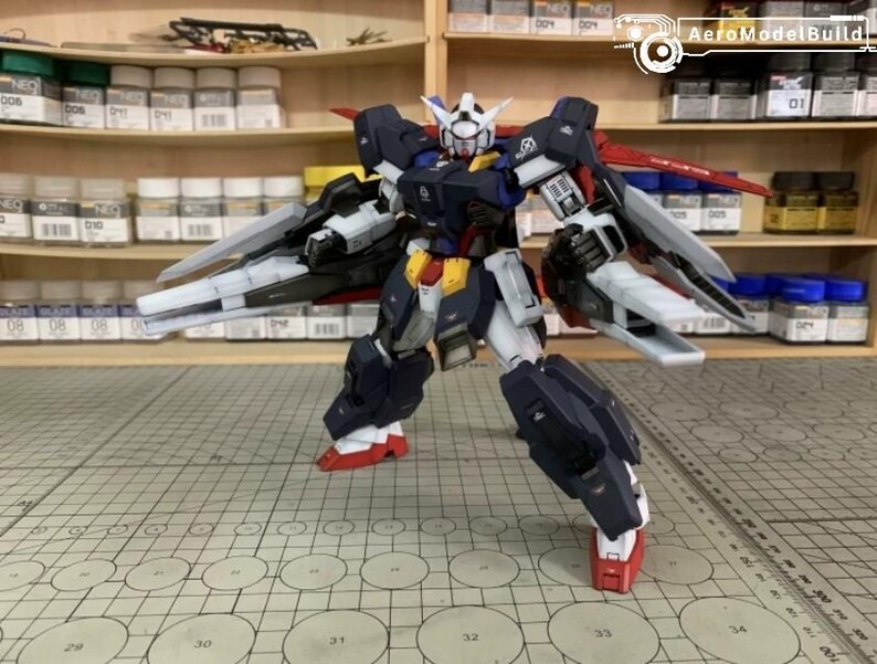 Puede incluir: Modelo detallado de un robot Gundam, con una combinaci&oacute;n de colores azul marino, blanco, rojo y amarillo. El robot tiene grandes alas y est&aacute; posado sobre una superficie cuadriculada. El fondo incluye estantes con botes de pintura y suministros de modelismo.