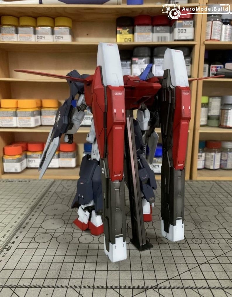 Puede incluir: Un kit de modelo de mecha parcialmente ensamblado, con componentes rojos, blancos y azul marino. El modelo tiene grandes piezas rectangulares para las piernas y alas. El kit est&aacute; sobre una esterilla de corte con un patr&oacute;n de cuadr&iacute;cula.