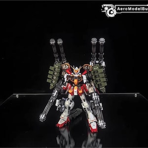 May include: A detailed model robot in red, white, orange, and grey. It features multiple large guns and green armour-like attachments. The AeroModelBuild logo is visible in the top right corner.