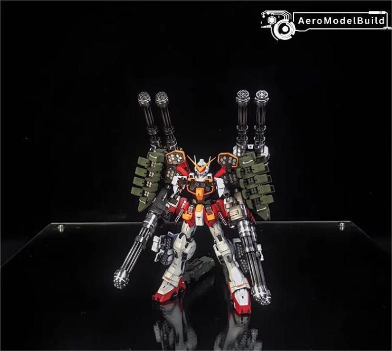 May include: A detailed model robot in red, white, orange, and grey. It features multiple large guns and green armour-like attachments. The AeroModelBuild logo is visible in the top right corner.
