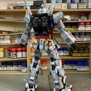 K&ouml;nnte beinhalten: Ein detailliertes Modell eines Gundam-Roboters, von hinten betrachtet. Das Modell hat ein Farbschema aus Wei&szlig;, Grau, Rot, Blau und Gelb. Der Roboter steht auf einer Rasteroberfl&auml;che, im Hintergrund Regale mit Farbe und Zubeh&ouml;r. Das Logo "AeroModelBuild" ist sichtbar.