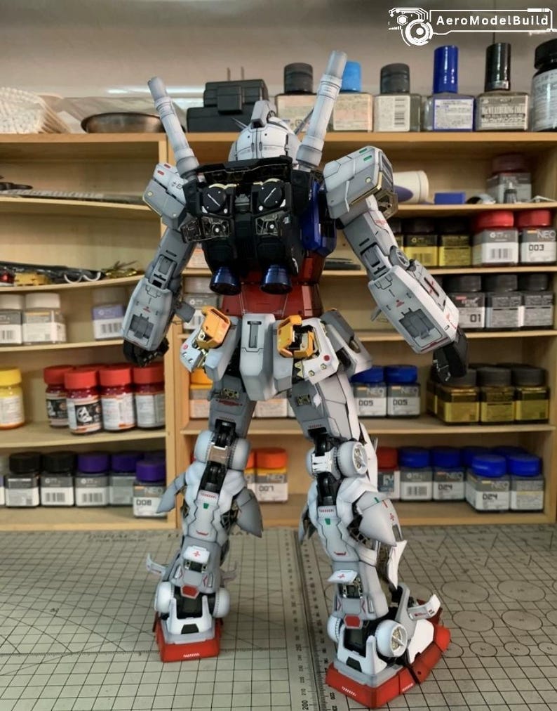 K&ouml;nnte beinhalten: Ein detailliertes Modell eines Gundam-Roboters, von hinten betrachtet. Das Modell hat ein Farbschema aus Wei&szlig;, Grau, Rot, Blau und Gelb. Der Roboter steht auf einer Rasteroberfl&auml;che, im Hintergrund Regale mit Farbe und Zubeh&ouml;r. Das Logo "AeroModelBuild" ist sichtbar.