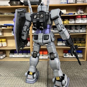 May include: A detailed model of a gray and purple robot with a shield and a gun. The robot has black accents and gold details. The model is standing on a cutting mat with a ruler. The text "ArrowModelBuild" is visible.