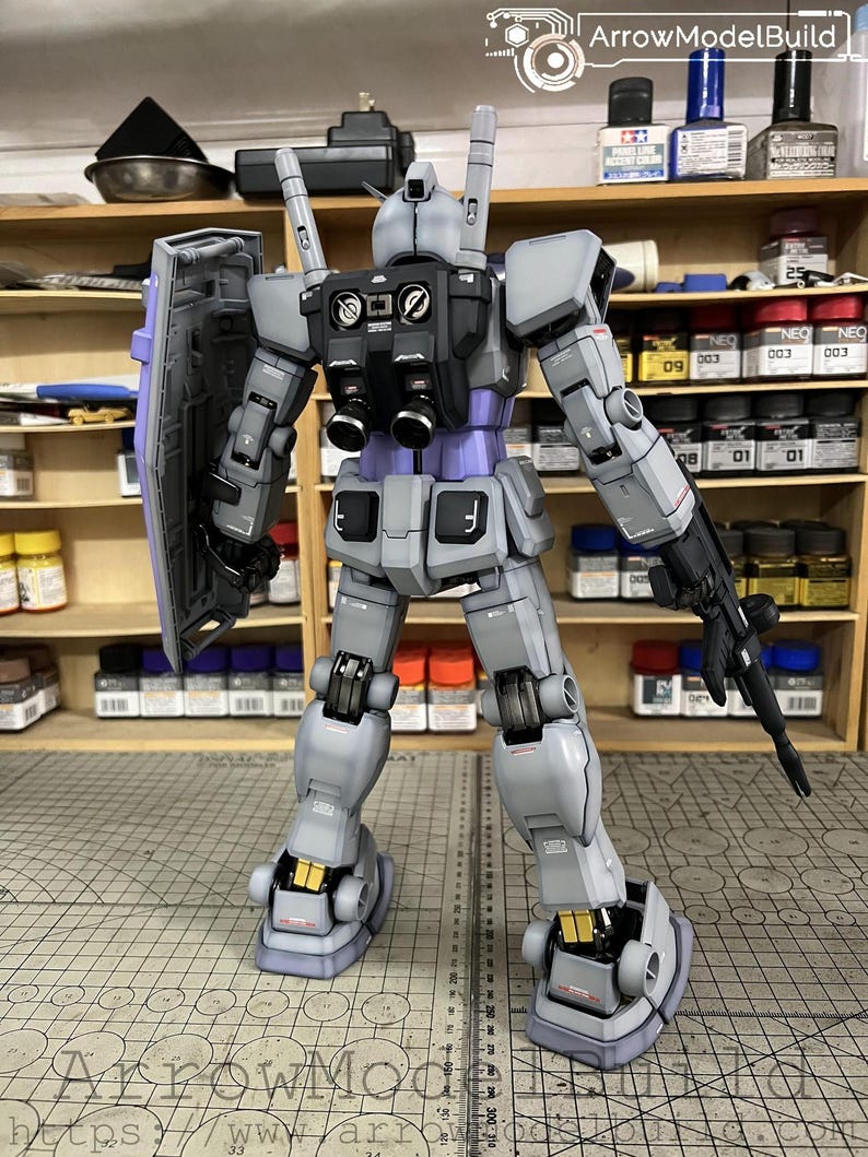 May include: A detailed model of a gray and purple robot with a shield and a gun. The robot has black accents and gold details. The model is standing on a cutting mat with a ruler. The text "ArrowModelBuild" is visible.