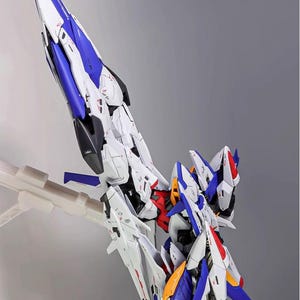 Puede incluir: Kit de modelo detallado de una aeronave o robot futurista, predominantemente blanco con detalles en azul, rojo y amarillo. El modelo est&aacute; montado sobre un soporte transparente sobre un fondo gris. El texto "AeroModelBuild" es visible.