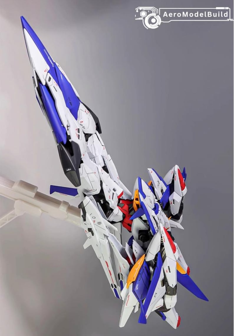 Puede incluir: Kit de modelo detallado de una aeronave o robot futurista, predominantemente blanco con detalles en azul, rojo y amarillo. El modelo est&aacute; montado sobre un soporte transparente sobre un fondo gris. El texto "AeroModelBuild" es visible.