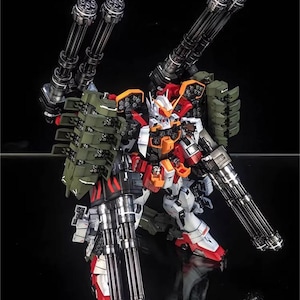 May include: A detailed model robot, predominantly white, red, and orange, with green detailing. It features multiple large, silver-coloured machine guns attached to its arms and back. The text "AeroModelBuild" is visible in the top right corner.