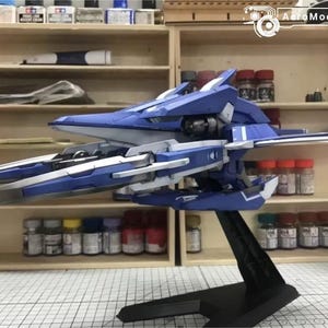 May include: A blue and white model airplane on a black stand. The model has detailed wings and a sleek design. The background includes shelves with paint bottles and other model-building supplies.