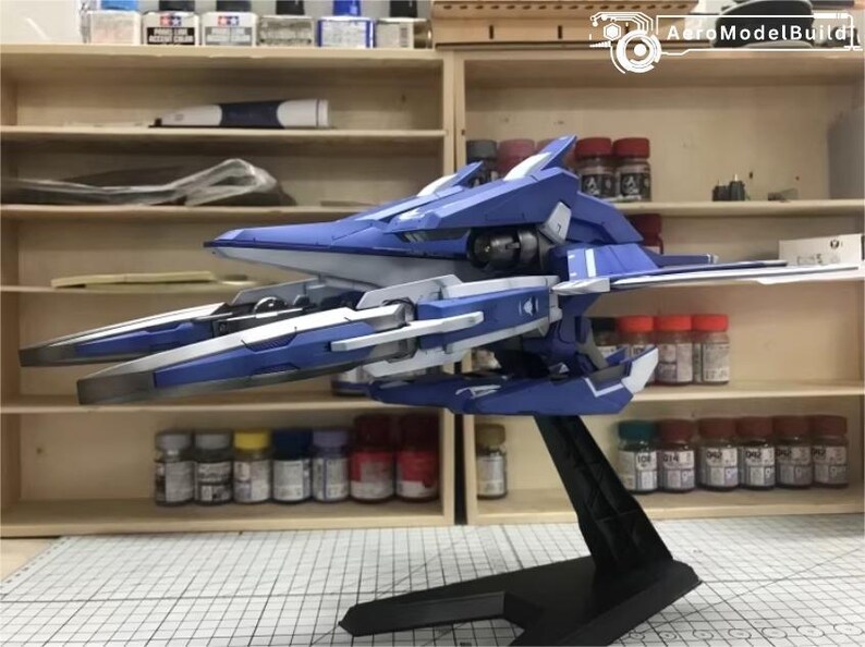 May include: A blue and white model airplane on a black stand. The model has detailed wings and a sleek design. The background includes shelves with paint bottles and other model-building supplies.