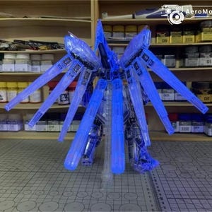 May include: A detailed blue and clear model robot kit, featuring large wing-like elements. The model is lit with blue light, highlighting its complex design. The background shows shelves stocked with model paints and tools, indicating a hobbyist's area.