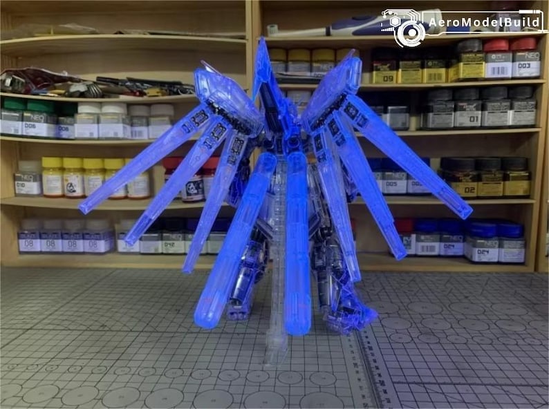 May include: A detailed blue and clear model robot kit, featuring large wing-like elements. The model is lit with blue light, highlighting its complex design. The background shows shelves stocked with model paints and tools, indicating a hobbyist's area.
