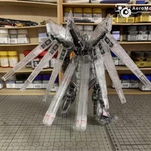 May include: A clear, unassembled robot model kit with large, wing-like structures. The model is made of transparent plastic, revealing internal details. The kit is on a grid-patterned surface, with shelves of paint and supplies behind it.