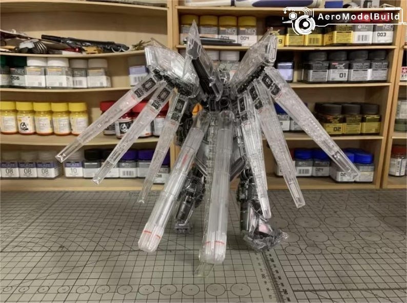 May include: A clear, unassembled robot model kit with large, wing-like structures. The model is made of transparent plastic, revealing internal details. The kit is on a grid-patterned surface, with shelves of paint and supplies behind it.