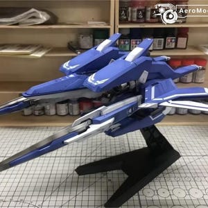 May include: A blue and white model airplane with a sleek design, featuring sharp angles and detailed wings. The model is mounted on a black stand, showcasing its aerodynamic form. The background includes shelves with paint supplies.