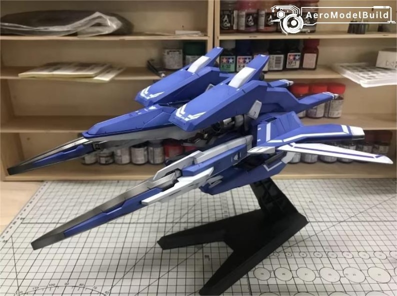 May include: A blue and white model airplane with a sleek design, featuring sharp angles and detailed wings. The model is mounted on a black stand, showcasing its aerodynamic form. The background includes shelves with paint supplies.