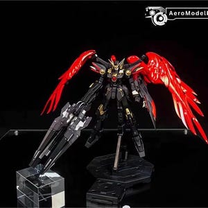 May include: A black and gold model robot with large red wings, displayed on a black stand. The robot has a detailed design with gold accents and is holding a large black weapon. The image includes the text "AeroModelBuild".