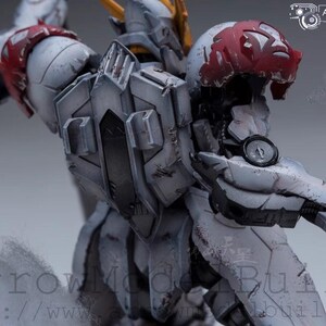 May include: A detailed, weathered model of a robotic figure, primarily in shades of gray and white, with red accents on the shoulder and head. The figure has intricate mechanical details and a circular gauge on its arm. The text "ArrowModelBuild" is visible.