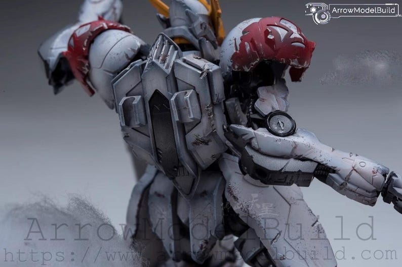 May include: A detailed, weathered model of a robotic figure, primarily in shades of gray and white, with red accents on the shoulder and head. The figure has intricate mechanical details and a circular gauge on its arm. The text "ArrowModelBuild" is visible.