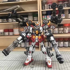 K&ouml;nnte beinhalten: Detailliertes Modell eines Gundam-Roboters mit wei&szlig;em, rotem, gelbem und schwarzem Farbschema. Der Roboter hat mehrere gro&szlig;e Waffen an Armen und Schultern. Das Modell steht auf einer Oberfl&auml;che mit Rastermuster.