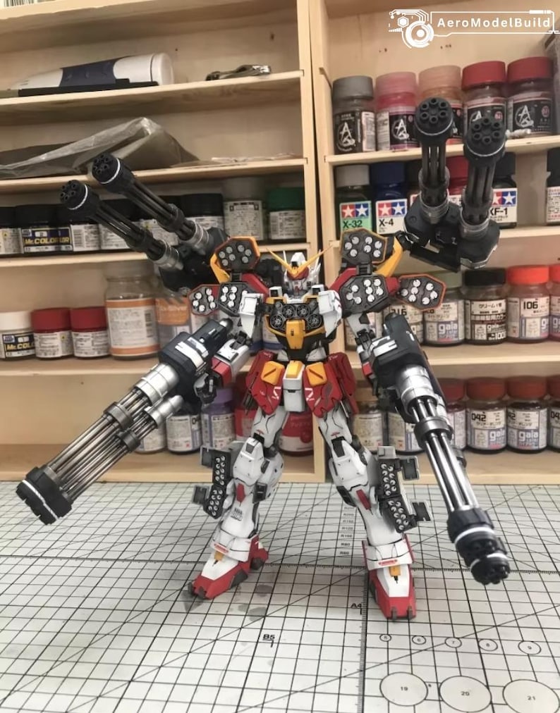 K&ouml;nnte beinhalten: Detailliertes Modell eines Gundam-Roboters mit wei&szlig;em, rotem, gelbem und schwarzem Farbschema. Der Roboter hat mehrere gro&szlig;e Waffen an Armen und Schultern. Das Modell steht auf einer Oberfl&auml;che mit Rastermuster.