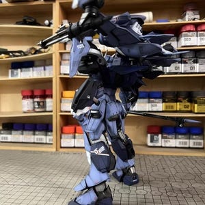 May include: A detailed model of a futuristic robot, primarily in shades of blue and black, with intricate mechanical details. The model is posed on a grid surface, showcasing its weaponry and design. The background includes shelves with paint supplies.