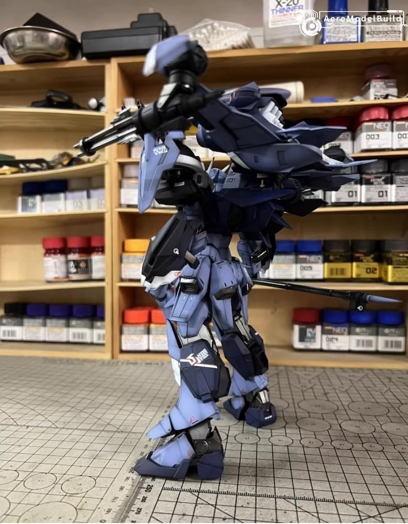 May include: A detailed model of a futuristic robot, primarily in shades of blue and black, with intricate mechanical details. The model is posed on a grid surface, showcasing its weaponry and design. The background includes shelves with paint supplies.