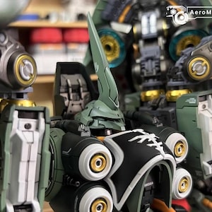 May include: Close-up of a detailed, assembled model kit of a mech robot. The robot features a green and black color scheme with gold accents. The head has a prominent, pointed crest. The AeroModelBuild logo is visible in the top right corner.