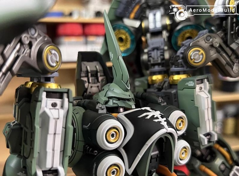 May include: Close-up of a detailed, assembled model kit of a mech robot. The robot features a green and black color scheme with gold accents. The head has a prominent, pointed crest. The AeroModelBuild logo is visible in the top right corner.