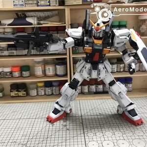 Op de afbeelding: Gedetailleerde modelbouwkit van een witte, rode en zwarte robot met een groot geweer en schild. De robot poseert voor een houten plank vol verfpotten en benodigdheden. Het model is ongeveer 30 cm hoog.