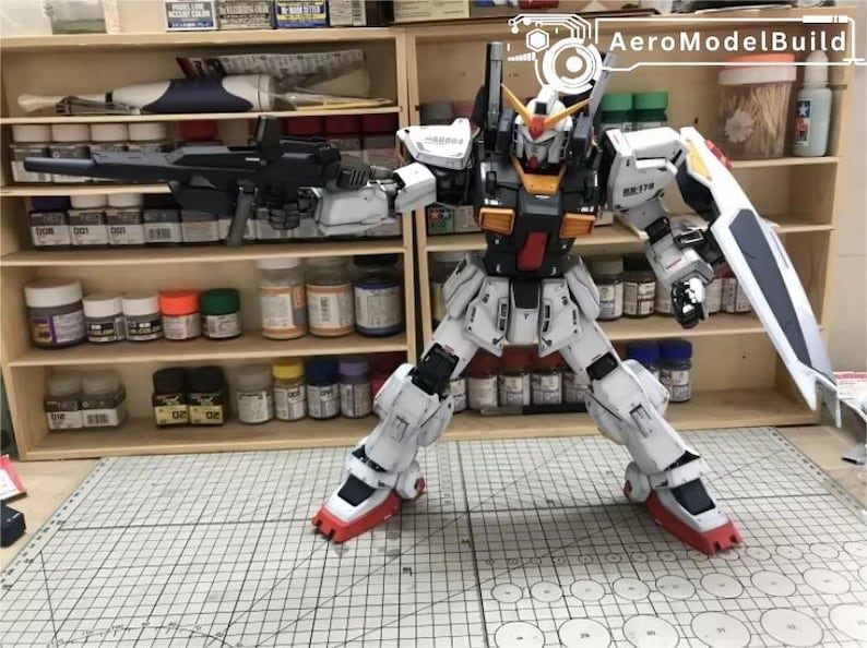 Op de afbeelding: Gedetailleerde modelbouwkit van een witte, rode en zwarte robot met een groot geweer en schild. De robot poseert voor een houten plank vol verfpotten en benodigdheden. Het model is ongeveer 30 cm hoog.