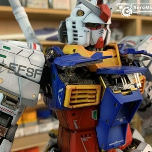K&ouml;nnte beinhalten: Detailliertes Modell eines Gundam-Roboters mit wei&szlig;em Kopf, roten und gelben Akzenten, blauem und gelbem Torso und roten Beinen. Das Modell weist komplizierte mechanische Details auf, und der Text "EFSF" ist auf der Schulter sichtbar.