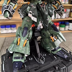 May include: A detailed, olive green and black model kit of a mech robot, with orange and white accents. The model is on a black display stand. The image shows the robot's legs, torso, and arms, with visible mechanical details and panel lines.