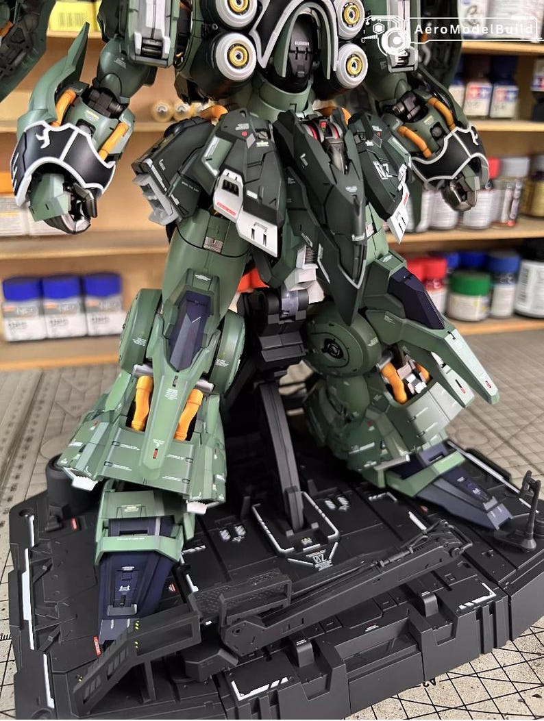 May include: A detailed, olive green and black model kit of a mech robot, with orange and white accents. The model is on a black display stand. The image shows the robot's legs, torso, and arms, with visible mechanical details and panel lines.