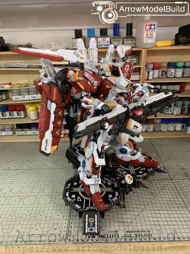 Puede incluir: Un detallado kit de modelo de un robot mecha, principalmente blanco y rojo, con intrincados detalles mec&aacute;nicos. El modelo est&aacute; montado sobre una plataforma en blanco y negro. La imagen incluye el texto "ArrowModelBuild" y "CUTTING MAT".