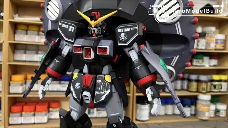 May include: A detailed model kit of a black, gray, and red robot with yellow accents. The robot has a large circular shield on its back and is labeled "GFAS-X1" and "DESTROY". The model is displayed in front of a shelf with various containers.
