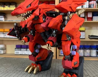 May include: A detailed, red and black model of a robotic dinosaur with gold-colored claws. The model has articulated joints and a menacing design, showcasing intricate details. The model is on a cutting mat with a grid pattern.