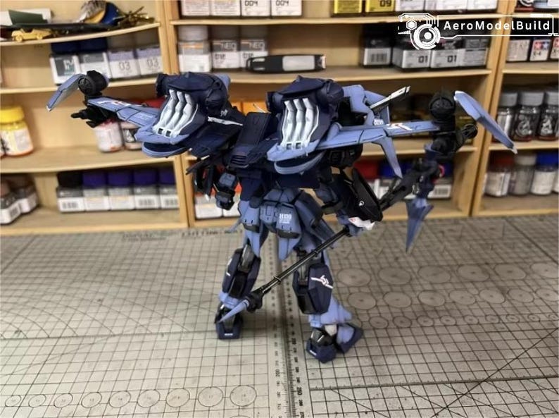 May include: A detailed model of a futuristic robot, primarily in shades of blue and black, with intricate mechanical details. The robot has large, wing-like structures and is holding a weapon. The model is set against a backdrop of shelves filled with model-building supplies.