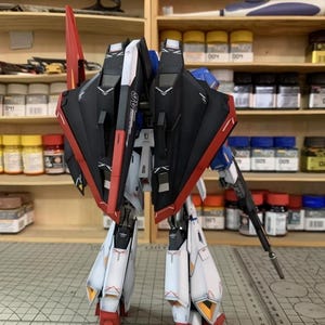 May include: A detailed model robot, predominantly red, black, white, and blue. The model features wings, legs, and a gun, showcasing intricate detailing. The background includes shelves stocked with paint bottles and tools, suggesting a hobbyist's workspace.