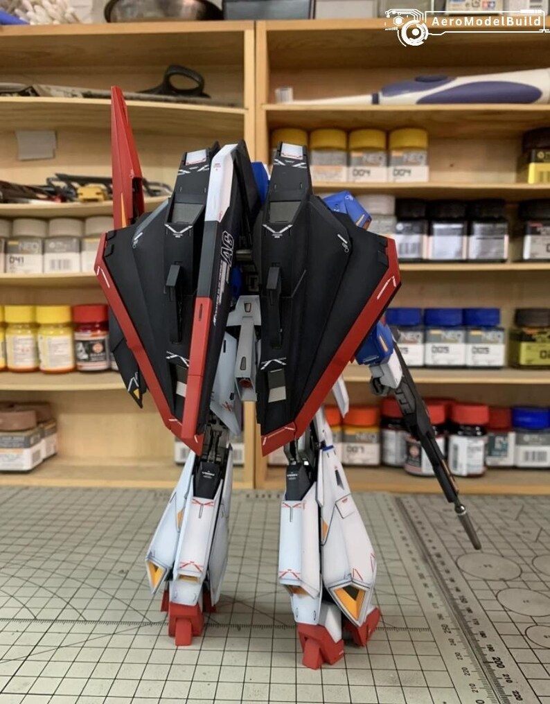 May include: A detailed model robot, predominantly red, black, white, and blue. The model features wings, legs, and a gun, showcasing intricate detailing. The background includes shelves stocked with paint bottles and tools, suggesting a hobbyist's workspace.