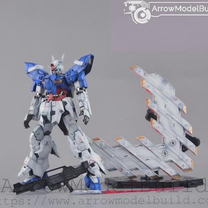 May include: A detailed model kit of a robot figure, mainly white, blue, and red. The robot is standing with a black weapon, and a separate, stylized wing-like structure is positioned nearby. The text "ArrowModelBuild" is visible.