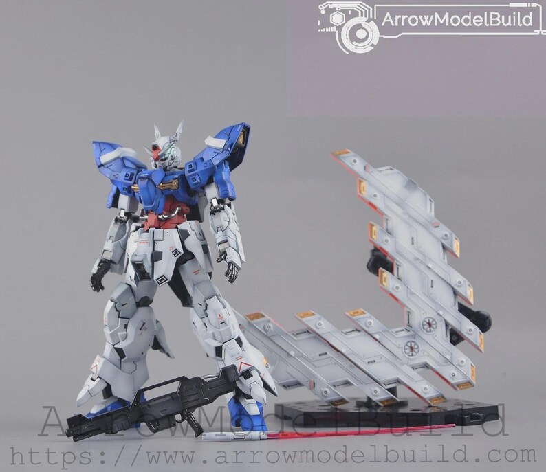 May include: A detailed model kit of a robot figure, mainly white, blue, and red. The robot is standing with a black weapon, and a separate, stylized wing-like structure is positioned nearby. The text "ArrowModelBuild" is visible.