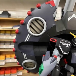 May include: Close-up of a detailed model robot with a dark gray shield featuring red accents and a circular vent. The model has the text "GFAS-X1" on a white and black label. The model is a complex build with multiple parts.