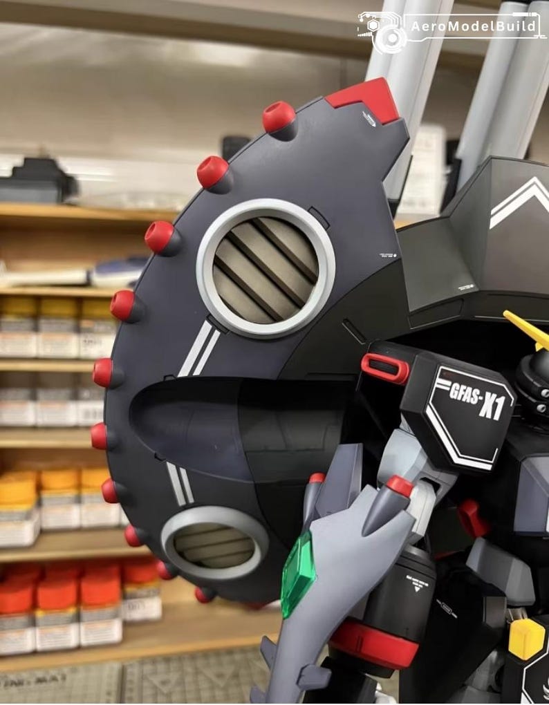 May include: Close-up of a detailed model robot with a dark gray shield featuring red accents and a circular vent. The model has the text "GFAS-X1" on a white and black label. The model is a complex build with multiple parts.