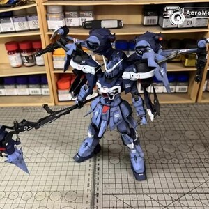 May include: A detailed model of a dark blue and white mecha robot with multiple weapons. The robot is standing on a grid surface, with shelves of paint and supplies in the background. The AeroModelBuild logo is visible in the top right corner.