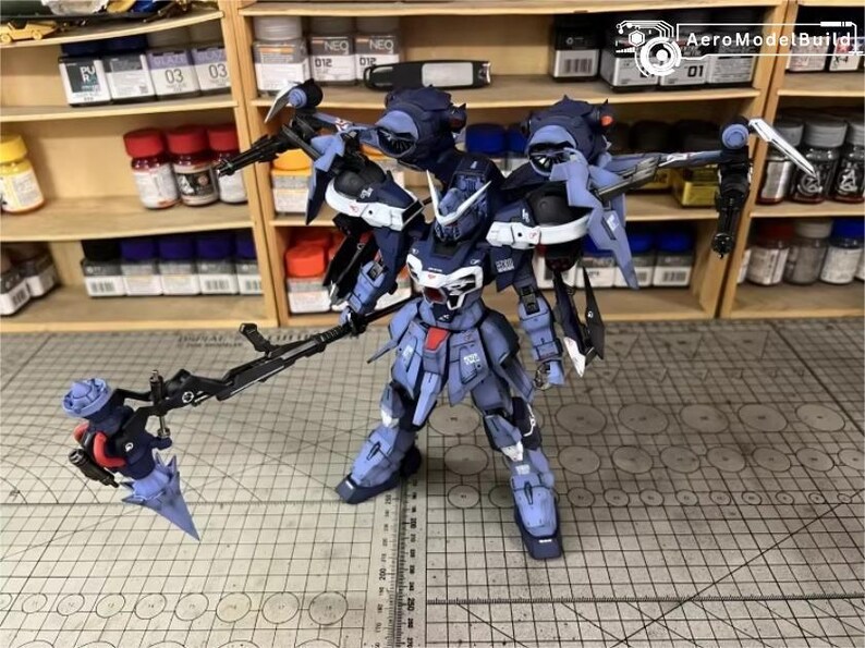 May include: A detailed model of a dark blue and white mecha robot with multiple weapons. The robot is standing on a grid surface, with shelves of paint and supplies in the background. The AeroModelBuild logo is visible in the top right corner.