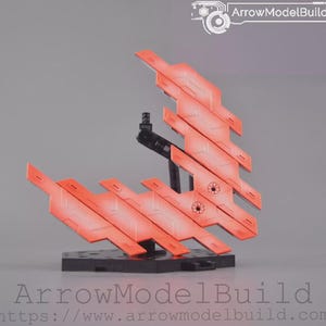 May include: A model kit component with orange and red angular panels, attached to a black stand and base. The panels have a gradient effect, transitioning from orange to red. The base is black, and the background is a solid grey.