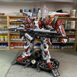 Puede incluir: Kit de modelo detallado de un robot mecha rojo, blanco y negro, completo con un rifle grande. El modelo est&aacute; posado sobre una plataforma negra, con varias herramientas y suministros visibles en el fondo. El texto "ArrowModelBuild" es visible.