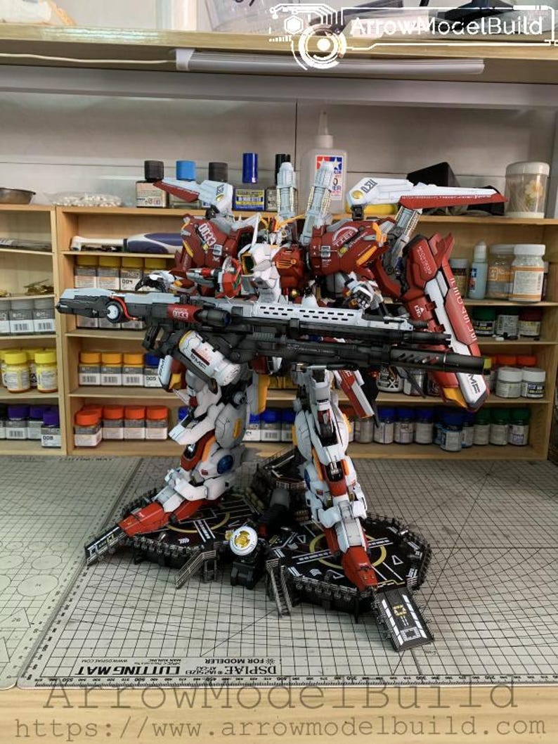 Puede incluir: Kit de modelo detallado de un robot mecha rojo, blanco y negro, completo con un rifle grande. El modelo est&aacute; posado sobre una plataforma negra, con varias herramientas y suministros visibles en el fondo. El texto "ArrowModelBuild" es visible.